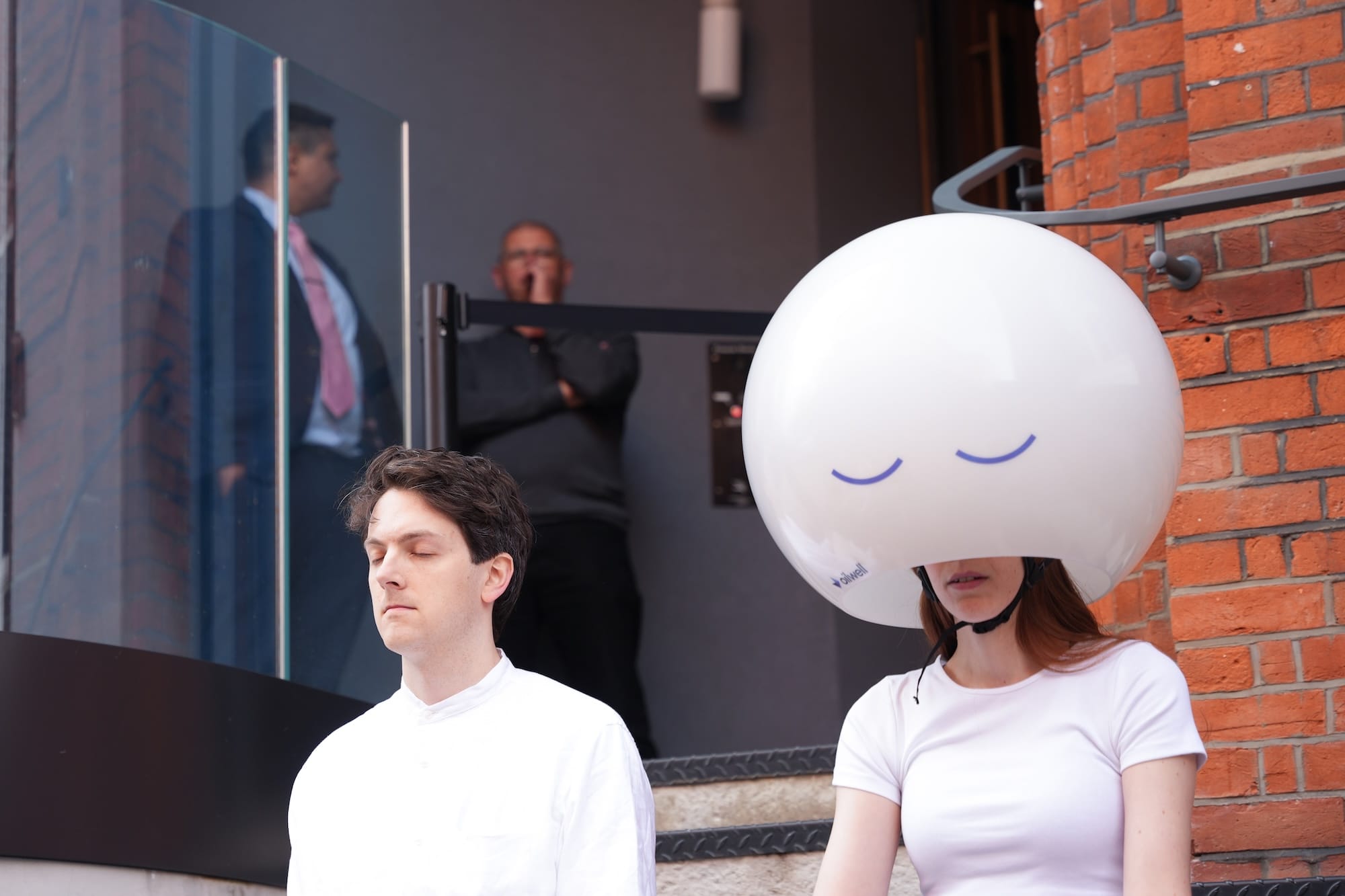 Close-up of two people in white clothing sitting in meditation poses on concrete steps. The person on the right wears a large white spherical helmet with a simple smiling face drawn on it, while the person on the left sits with eyes closed in a peaceful meditative state.